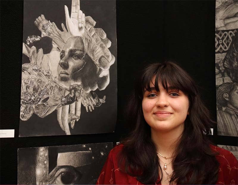 A student stands in front of her elaborate pencil drawings